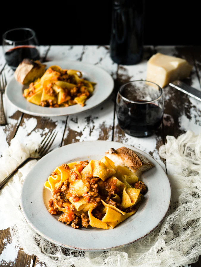 Two white dinner plates with pasta sitting next to glasses of red wine.