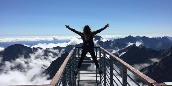 Person Standing on Hand Rails With Arms Wide Open Facing the Mountains and Clouds