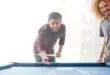 A group of four friends plays billiards on a pool table with blue felt in someone's home. There's a large sliding door in the background.