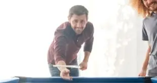 A group of four friends plays billiards on a pool table with blue felt in someone's home. There's a large sliding door in the background.