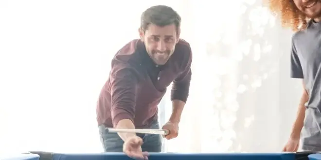 A group of four friends plays billiards on a pool table with blue felt in someone's home. There's a large sliding door in the background.