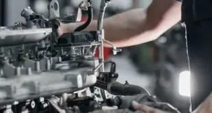 Metal components of a vehicle's engine are visible as a mechanic conducts repairs A white light brightens the workspace.