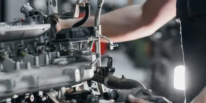 Metal components of a vehicle's engine are visible as a mechanic conducts repairs A white light brightens the workspace.