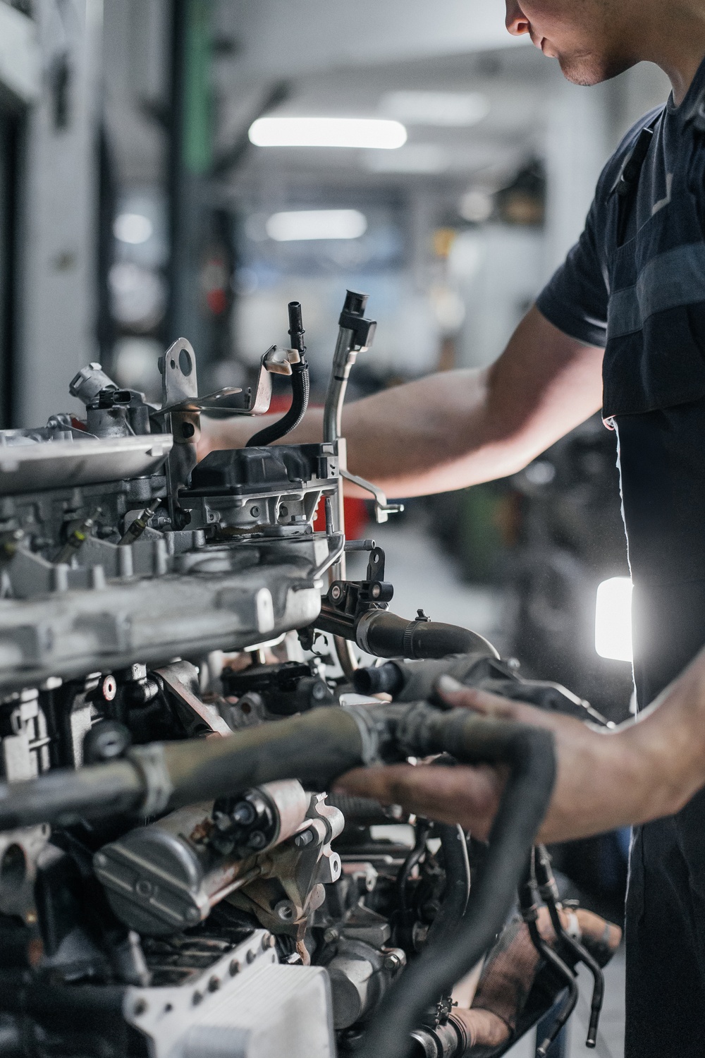 Metal components of a vehicle's engine are visible as a mechanic conducts repairs A white light brightens the workspace.