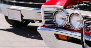 Bright classic cars line a street, with a red vintage car in front showing chrome headlights and a grille in sharp focus.