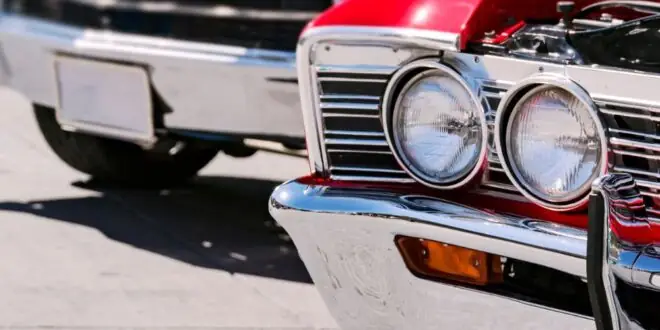 Bright classic cars line a street, with a red vintage car in front showing chrome headlights and a grille in sharp focus.
