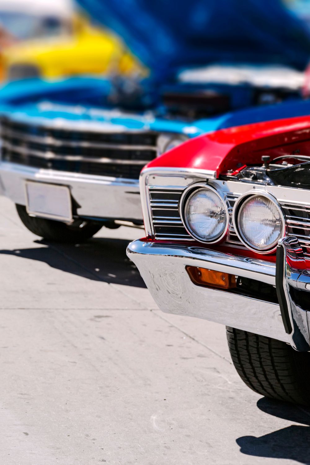 Bright classic cars line a street, with a red vintage car in front showing chrome headlights and a grille in sharp focus.