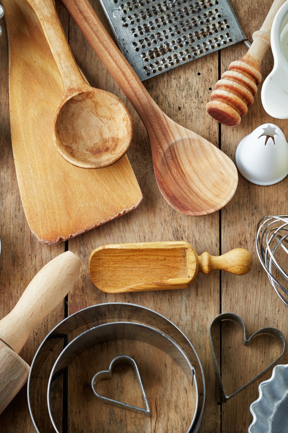A variety of kitchen tools, including spoons, a whisk, cutters, and a rolling pin, are spread across a rustic table.