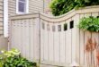 A wooden privacy fence frames a small patio garden with leafy shrubs, red plants, and a trellis by the house.