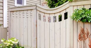 A wooden privacy fence frames a small patio garden with leafy shrubs, red plants, and a trellis by the house.