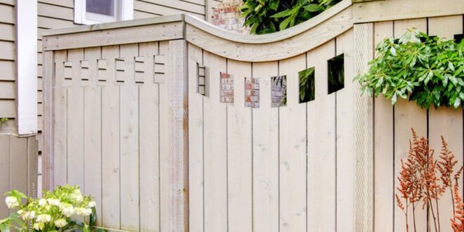 A wooden privacy fence frames a small patio garden with leafy shrubs, red plants, and a trellis by the house.