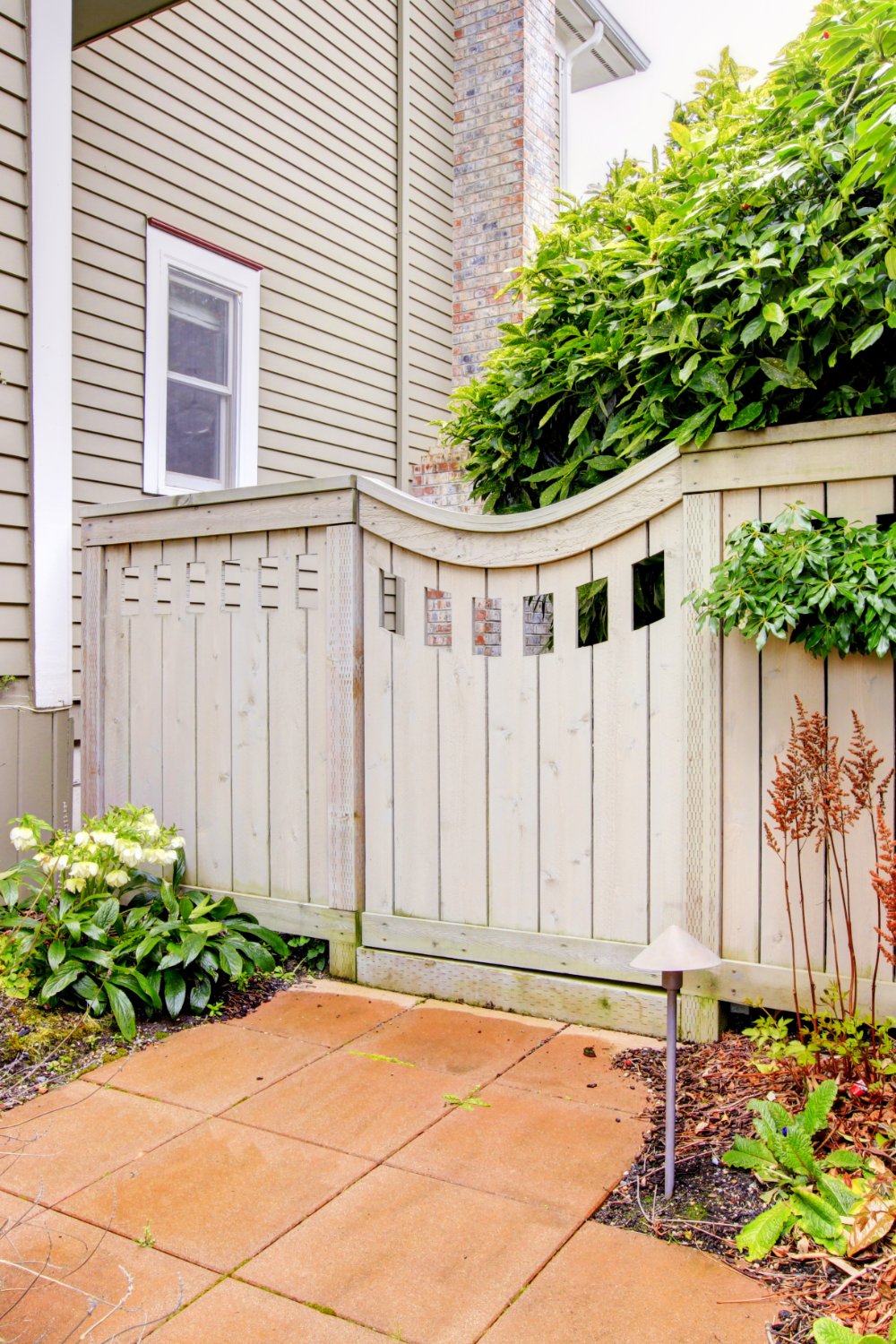 A wooden privacy fence frames a small patio garden with leafy shrubs, red plants, and a trellis by the house.
