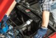 An overhead view of a man with a blue hat, gloves, and a flannel shirt working under the hood of an old red car.