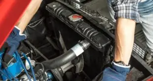 An overhead view of a man with a blue hat, gloves, and a flannel shirt working under the hood of an old red car.