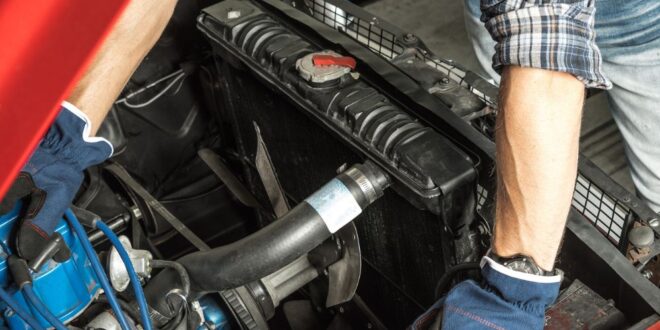 An overhead view of a man with a blue hat, gloves, and a flannel shirt working under the hood of an old red car.