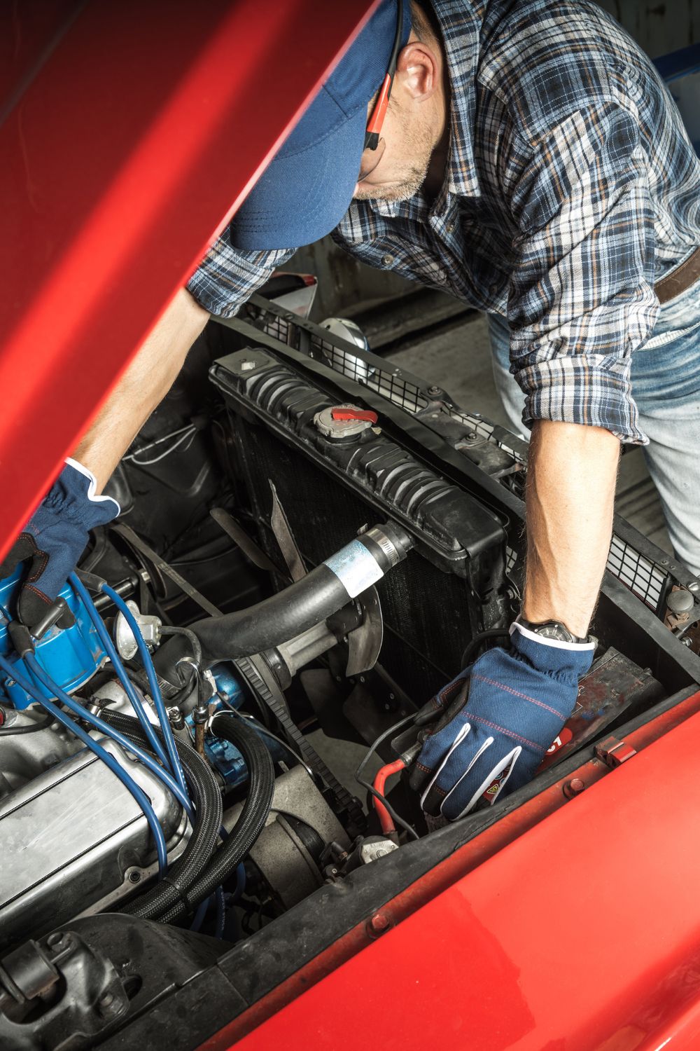 An overhead view of a man with a blue hat, gloves, and a flannel shirt working under the hood of an old red car.