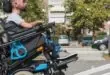 A woman helps a man in a wheelchair use the ramp in the back of an accessibility van. They are in a parking lot.
