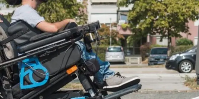 A woman helps a man in a wheelchair use the ramp in the back of an accessibility van. They are in a parking lot.