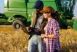 Two people standing in a wheat field and looking at a laptop, with a green combine harvester behind them.