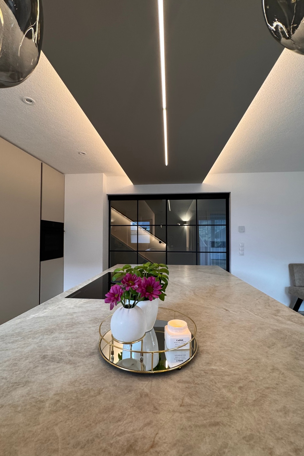 A long kitchen island has a brown and gray stone countertop. A circular tray with floral vases is on the counter.