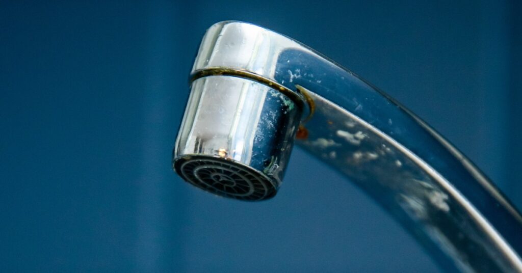 A close-up of a chrome faucet releasing tiny drops of water. Square, dark blue tiles fill the background.