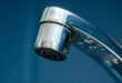 A close-up of a chrome faucet releasing tiny drops of water. Square, dark blue tiles fill the background.