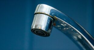 A close-up of a chrome faucet releasing tiny drops of water. Square, dark blue tiles fill the background.