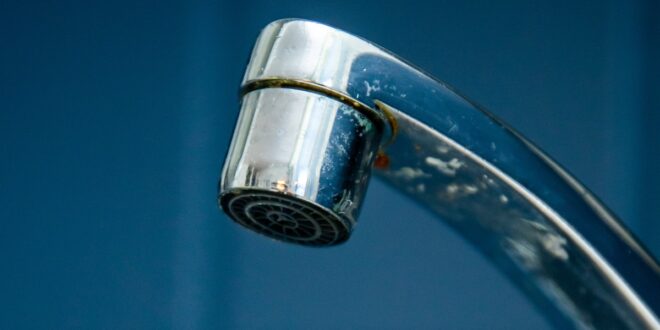 A close-up of a chrome faucet releasing tiny drops of water. Square, dark blue tiles fill the background.