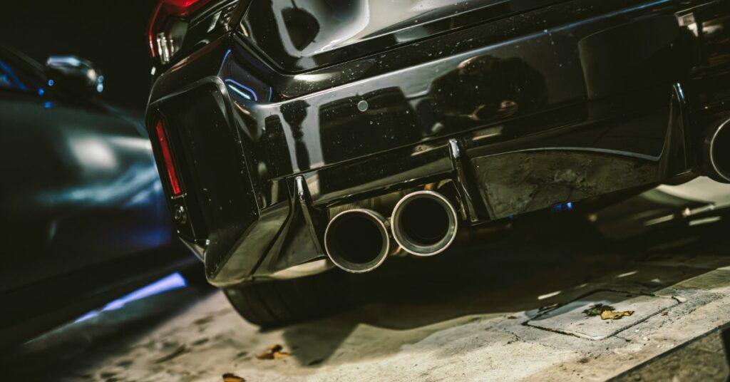 A black sports car rear bumper shows four round exhaust pipes, red taillights, and a large wing in a garage.