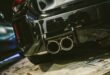 A black sports car rear bumper shows four round exhaust pipes, red taillights, and a large wing in a garage.