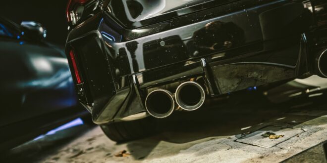 A black sports car rear bumper shows four round exhaust pipes, red taillights, and a large wing in a garage.