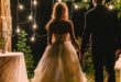 A bride in a white gown holding hands with a groom in a dark suit beneath hanging bulbs beside white lanterns at night.