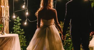 A bride in a white gown holding hands with a groom in a dark suit beneath hanging bulbs beside white lanterns at night.