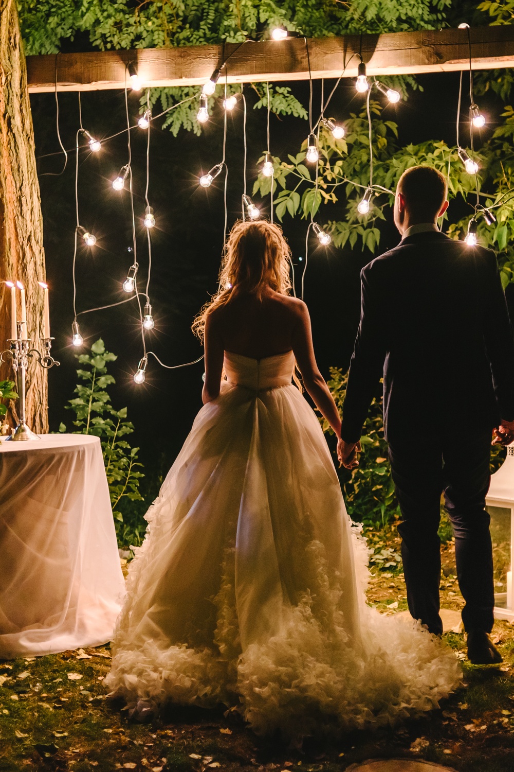 A bride in a white gown holding hands with a groom in a dark suit beneath hanging bulbs beside white lanterns at night.