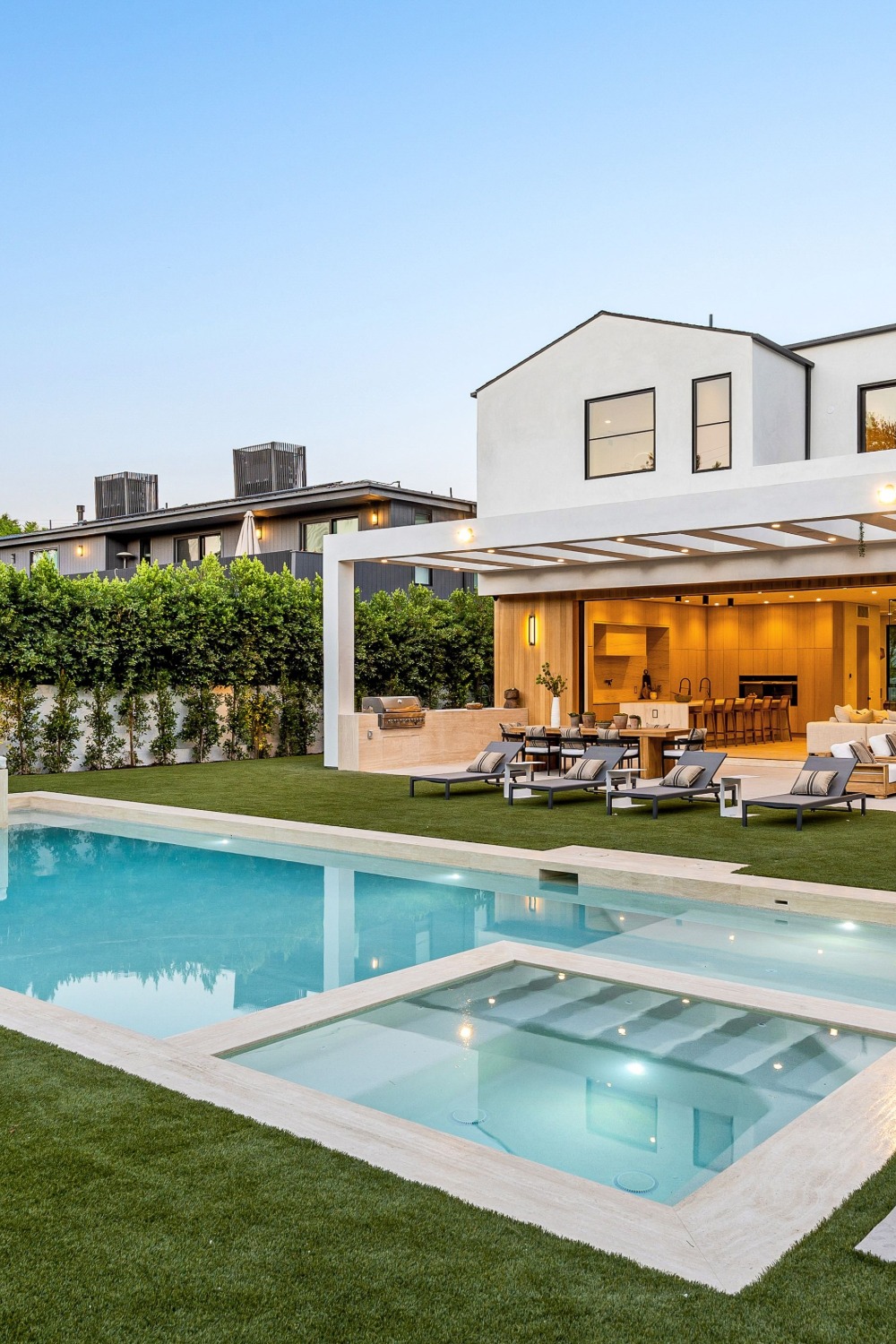 A modern white two-story home with a backyard swimming pool, raised spa, green lawn, and covered outdoor seating area.