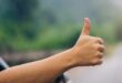 A close-up of a woman's hand sticking out of the window of a car with her thumb up and the road blurred in the background.