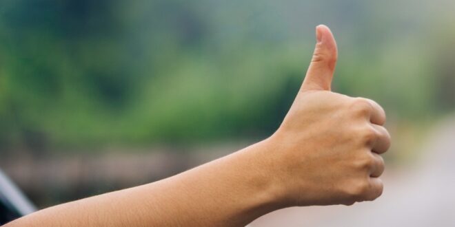 A close-up of a woman's hand sticking out of the window of a car with her thumb up and the road blurred in the background.