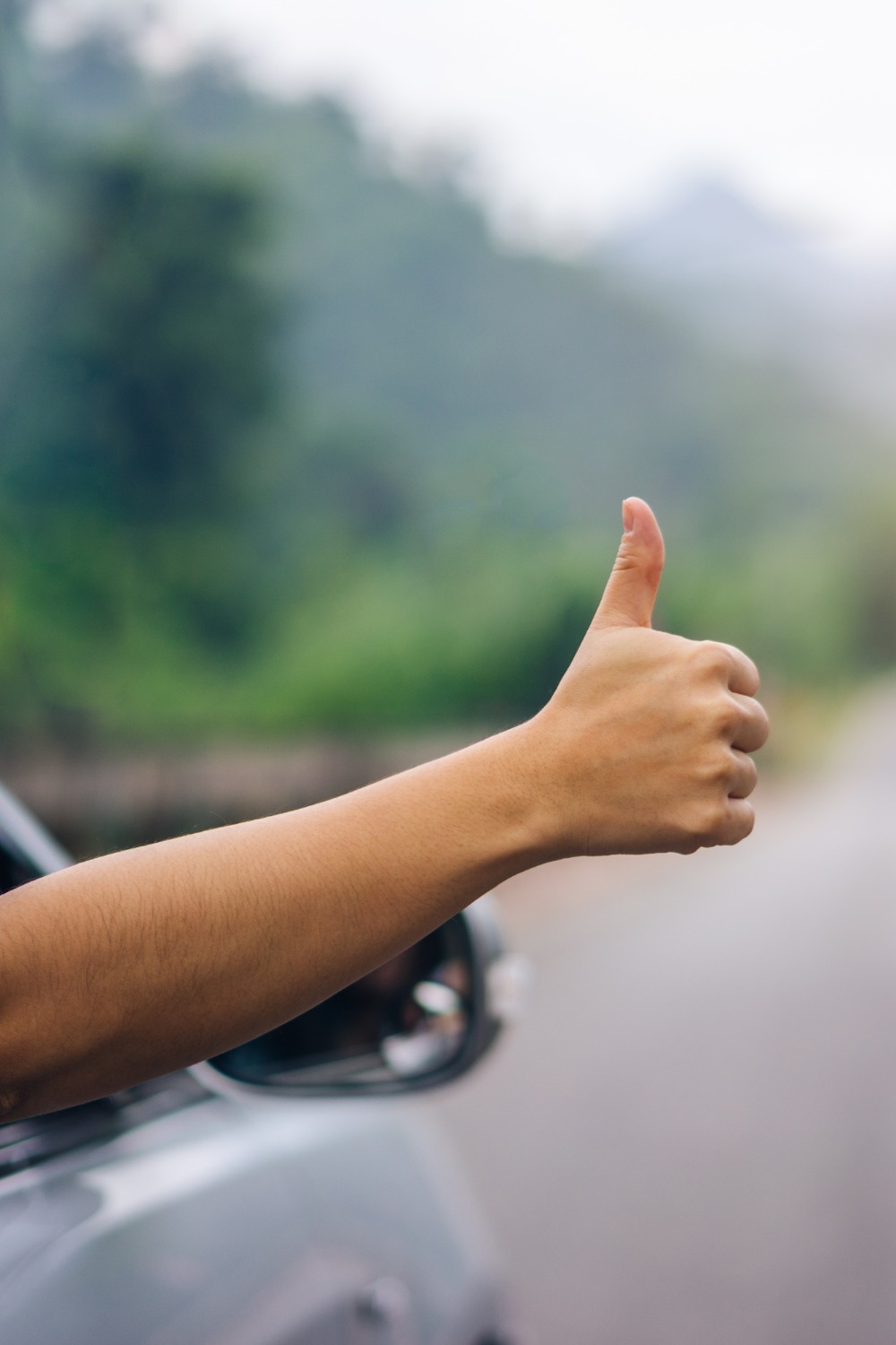 A close-up of a woman's hand sticking out of the window of a car with her thumb up and the road blurred in the background.